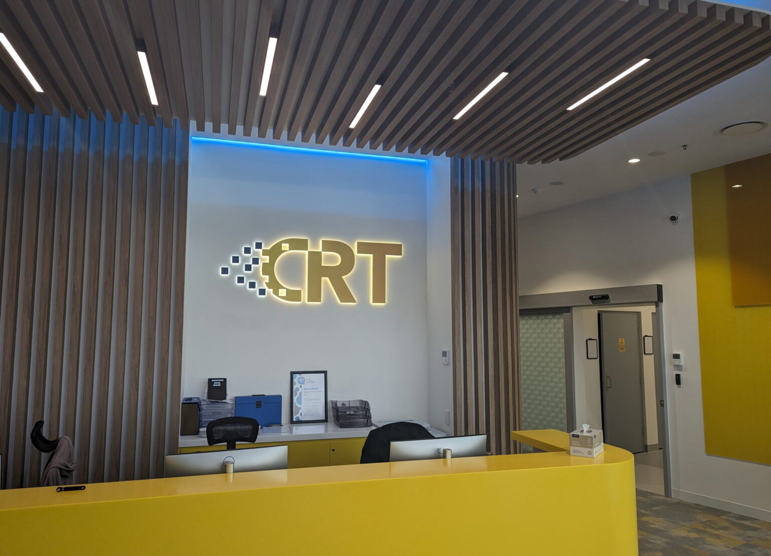 Reception area with a yellow curved counter and a glowing CRT logo on a white wall, framed by vertical wood slats.