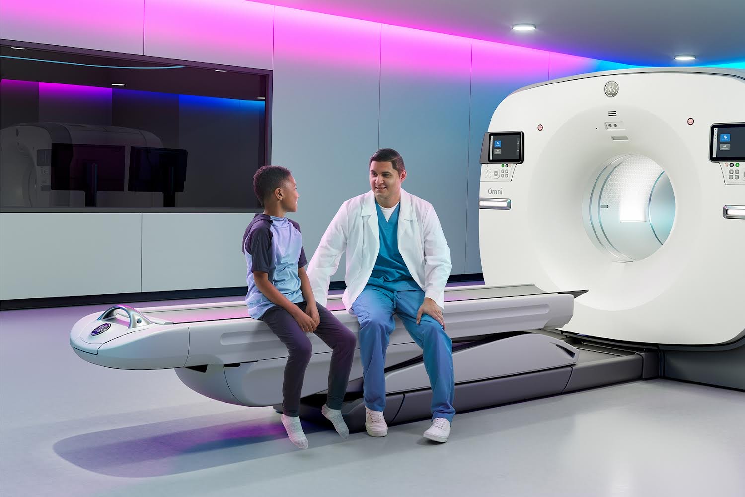 Doctor in scrubs and white coat sits with a young patient on an MRI scan bed in a bright imaging room with colorful lighting on the walls and ceiling.